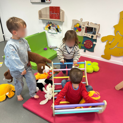 Trois jeunes enfants explorent une petite structure d'escalade en bois et jouent avec des peluches lors d'un atelier de jeux libres au Relais Petite Enfance.