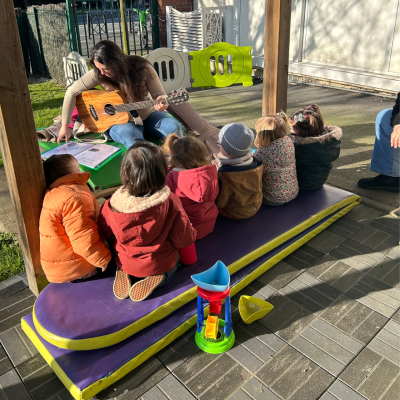 Une professionnelle joue de la guitare pour un groupe de jeunes enfants assis sur un tapis lors d'un atelier d'éveil musical en extérieur à la crèche L'île aux Enfants.