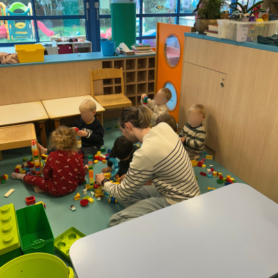 Un professionnel de la petite enfance et un groupe de jeunes enfants assis au sol jouent ensemble avec des blocs de construction colorés à la crèche Station Bout'chou.