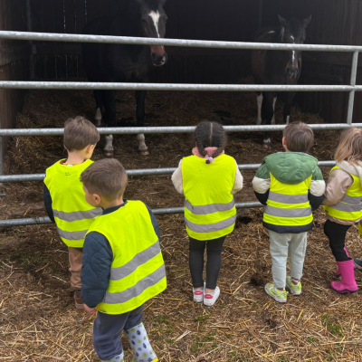 Cinq jeunes enfants en gilets jaunes de sécurité observent deux chevaux dans une étable lors d'une sortie à la ferme.